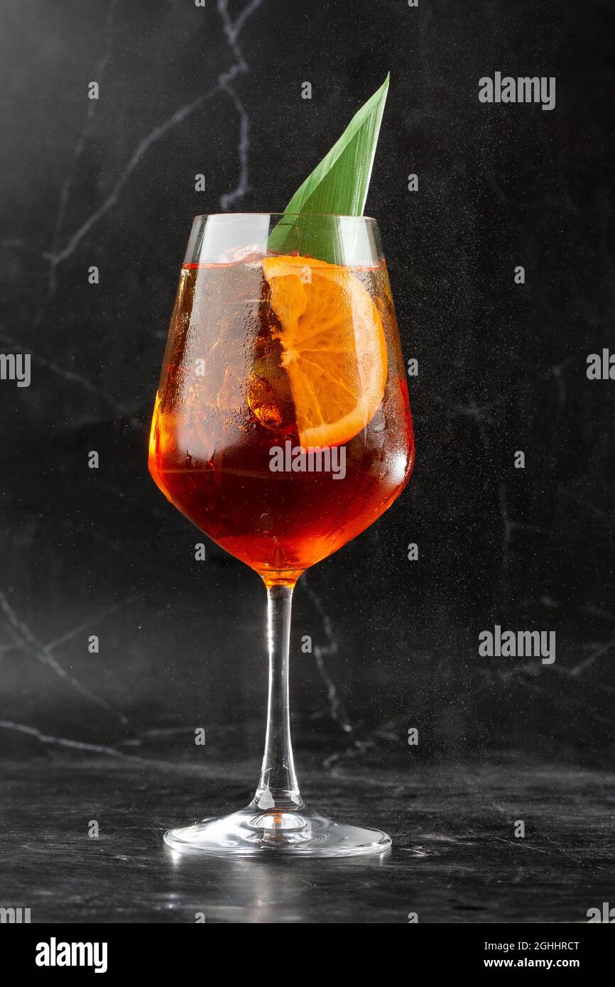 Ein Glas eisgekühlter Aperol Spritz-Cocktail, serviert in einem Weinglas, verziert mit Scheiben aus Orange und Basilikum, auf einem schwarzen Glas platziert Stockfoto