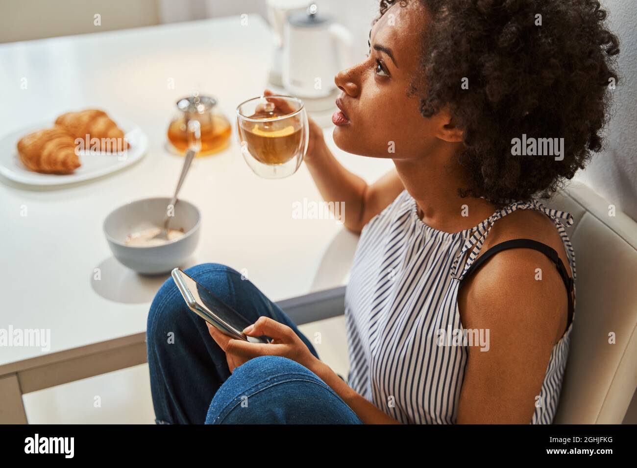 Frau mit einem Getränk und dem Handy am Küchentisch Stockfoto