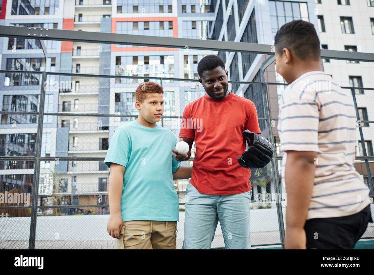 Inhalt African-American Coach in Handschuhen hält den Ball und erklärt Spielregeln, während er Jungen auf das Baseballspiel vorbereitet Stockfoto