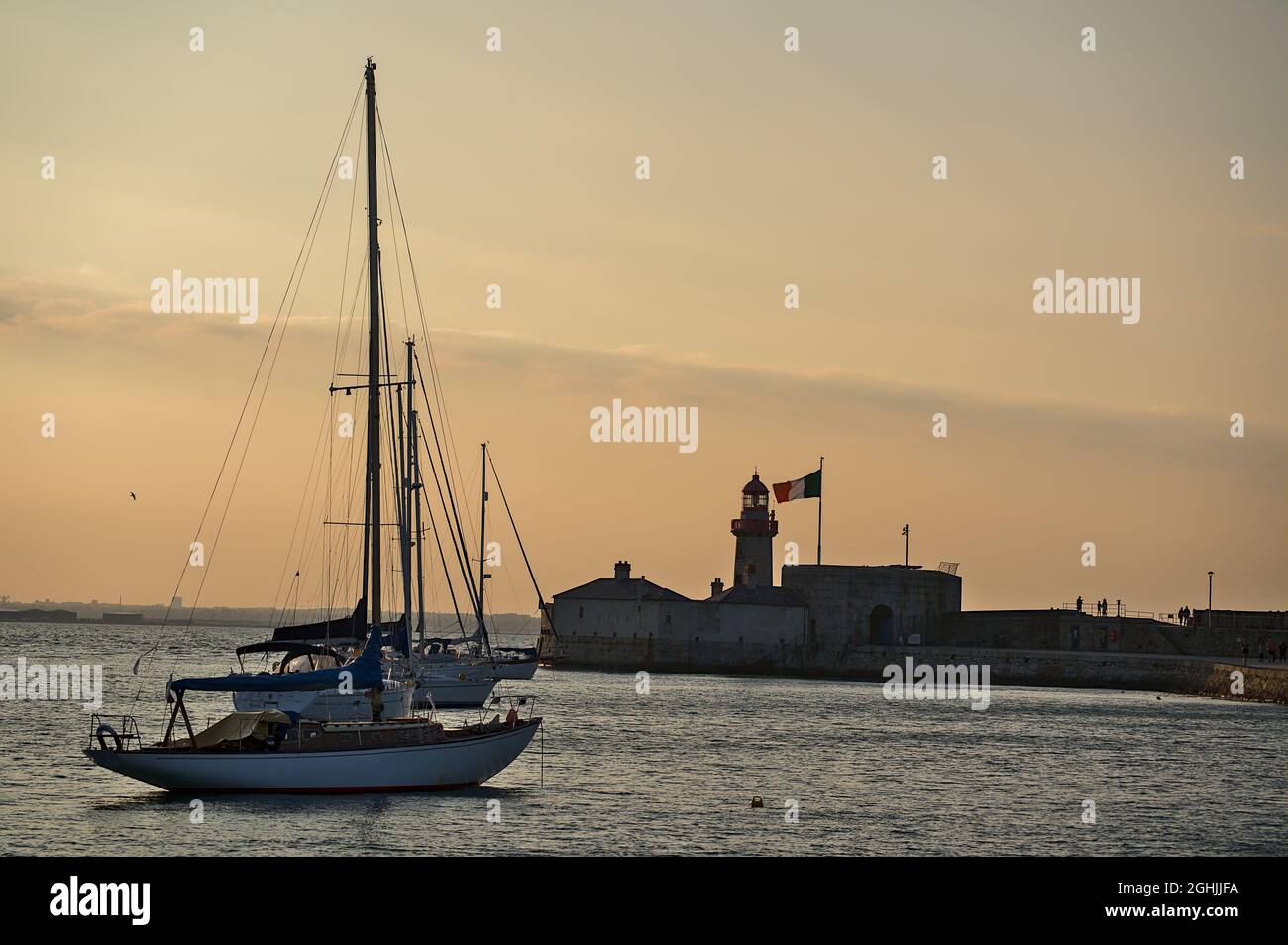 Dublin, Irland - 21. Juni 2021: Wunderschöne Abendansicht von Segelbooten, die in der Nähe des roten Leuchtturms am East Pier mit irischer Flagge im Hafen von Dun Laoghaire anlegen Stockfoto