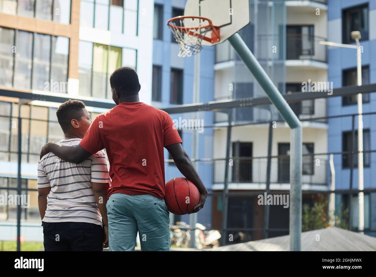 Rückansicht des schwarzen Vaters in orangefarbenem T-Shirt, der die Schulter des Sohnes umarmt, während er gegen den Basketballkorb steht Stockfoto