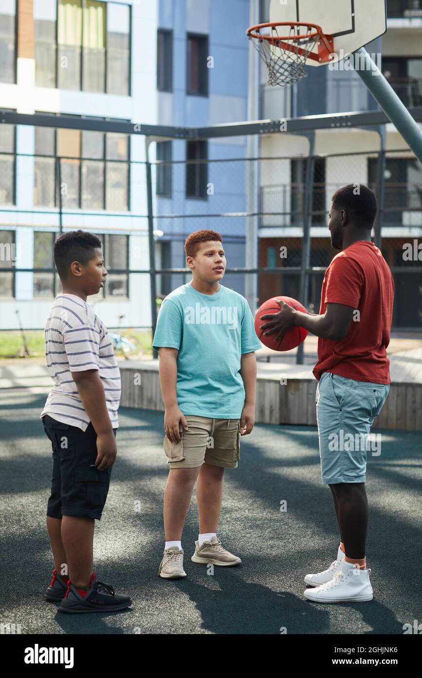 Afroamerikanischer Vater mit Basketball im Gespräch mit Jungen auf dem Spielplatz gegen modernes Gebäude Stockfoto