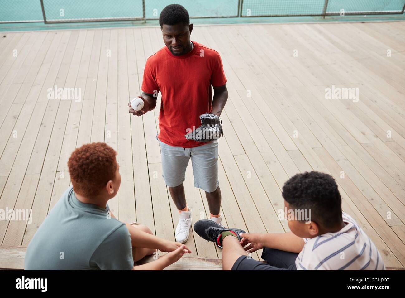 Junger afroamerikanischer Trainer in orangefarbenem T-Shirt hält Ball und Baseballhandschuh und erklärt Jungen die Pitching-Technik Stockfoto
