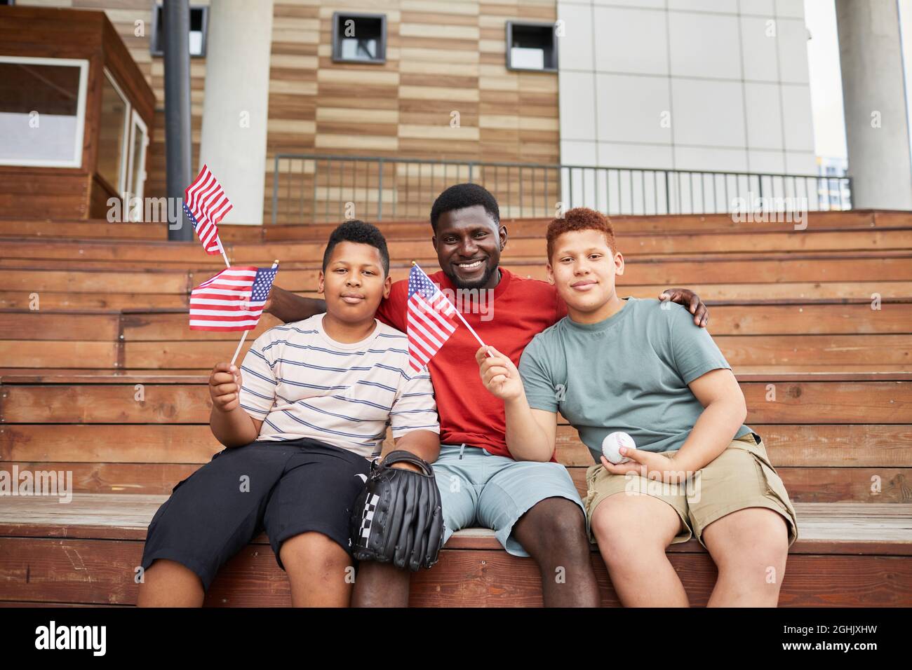Porträt eines glücklichen schwarzen Vaters, der Söhne im Teenageralter mit amerikanischen Flaggen umarmt, während sie gemeinsam das Baseballspiel besuchen Stockfoto