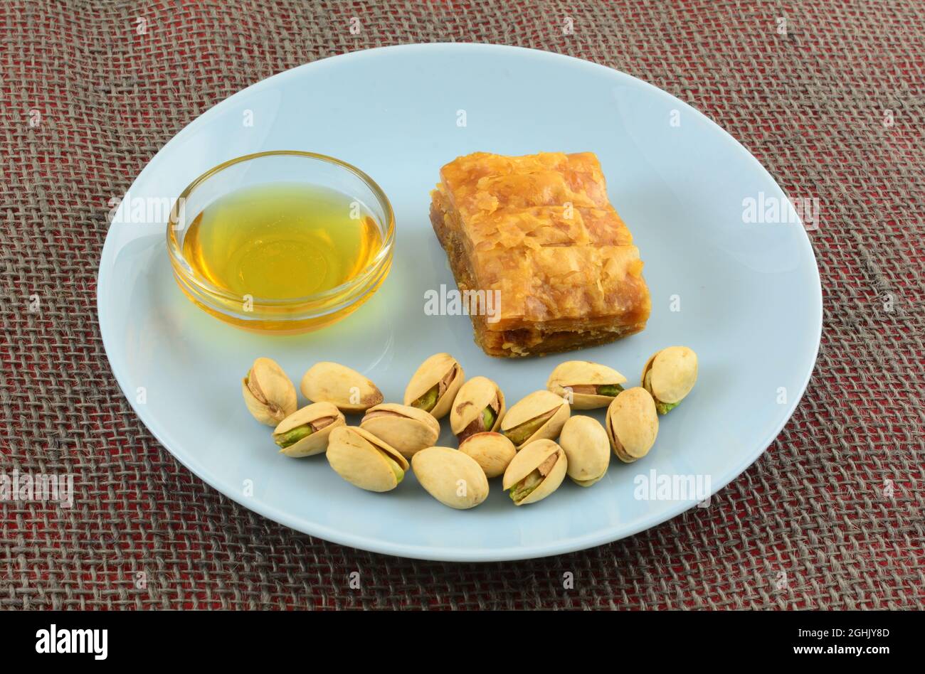 Stück Baklava mit Honig in Glasschale und Pistazien in Schalen auf blauem Snackteller Stockfoto