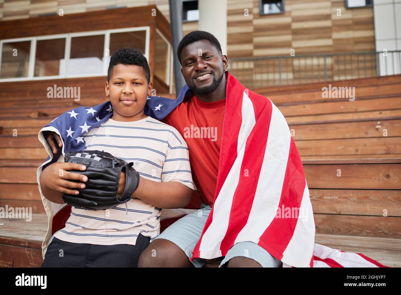 Porträt eines glücklichen schwarzen Vaters mittleren Alters, der unter amerikanischer Flagge sitzt und seinen Sohn in Baseballhandschuhen umarmt Stockfoto