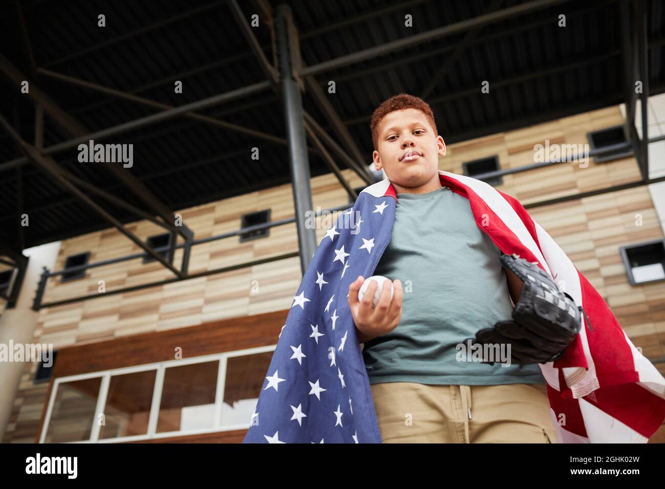 Unten Ansicht des selbstbewussten schwarzen Jungen mit amerikanischer Flagge bedeckt, der mit Baseballhandschuh und Ball unter dem Dach steht Stockfoto