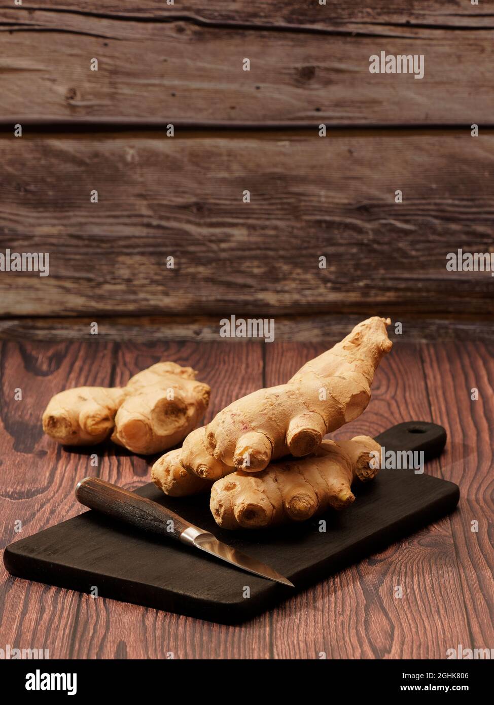 Ingwerwurzel und ein Messer auf einem verbrannten Holzschneidebrett auf braunem Holzhintergrund Stockfoto
