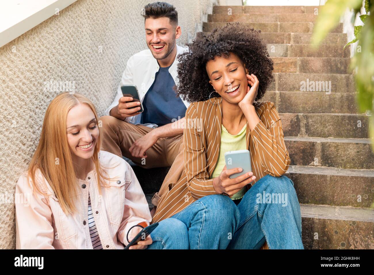 Fröhlicher, multirassischer junger Mann und Frauen in legerer Kleidung, lächelnd und mit Freude auf Handys surfen, während sie auf der Straße auf der Treppe sitzen Stockfoto