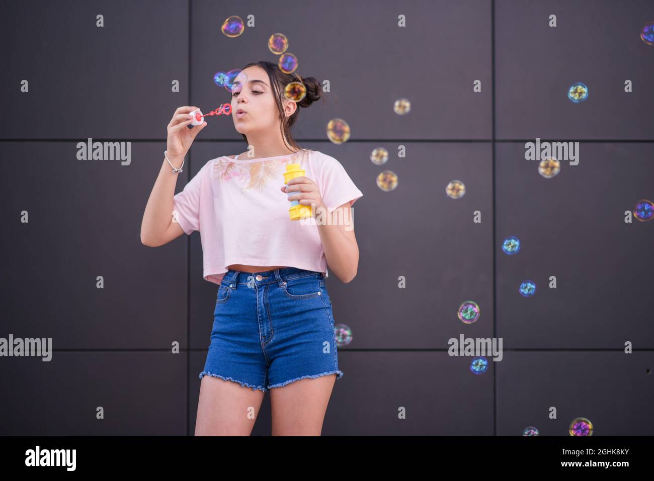 Unbeschwerte weibliche Teenager im trendigen lässigen Sommer-Outfit blasen bunte Seifenblasen gegen graue Wand Stockfoto