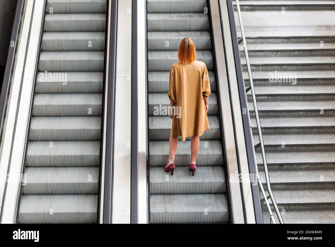 Ganzkörper-Rückansicht einer nicht erkennbaren Frau in stilvollem Kleid und hohen Absätzen, die allein auf einer Treppe der unterirdischen Rolltreppe stehen, die sich nach oben bewegt Stockfoto