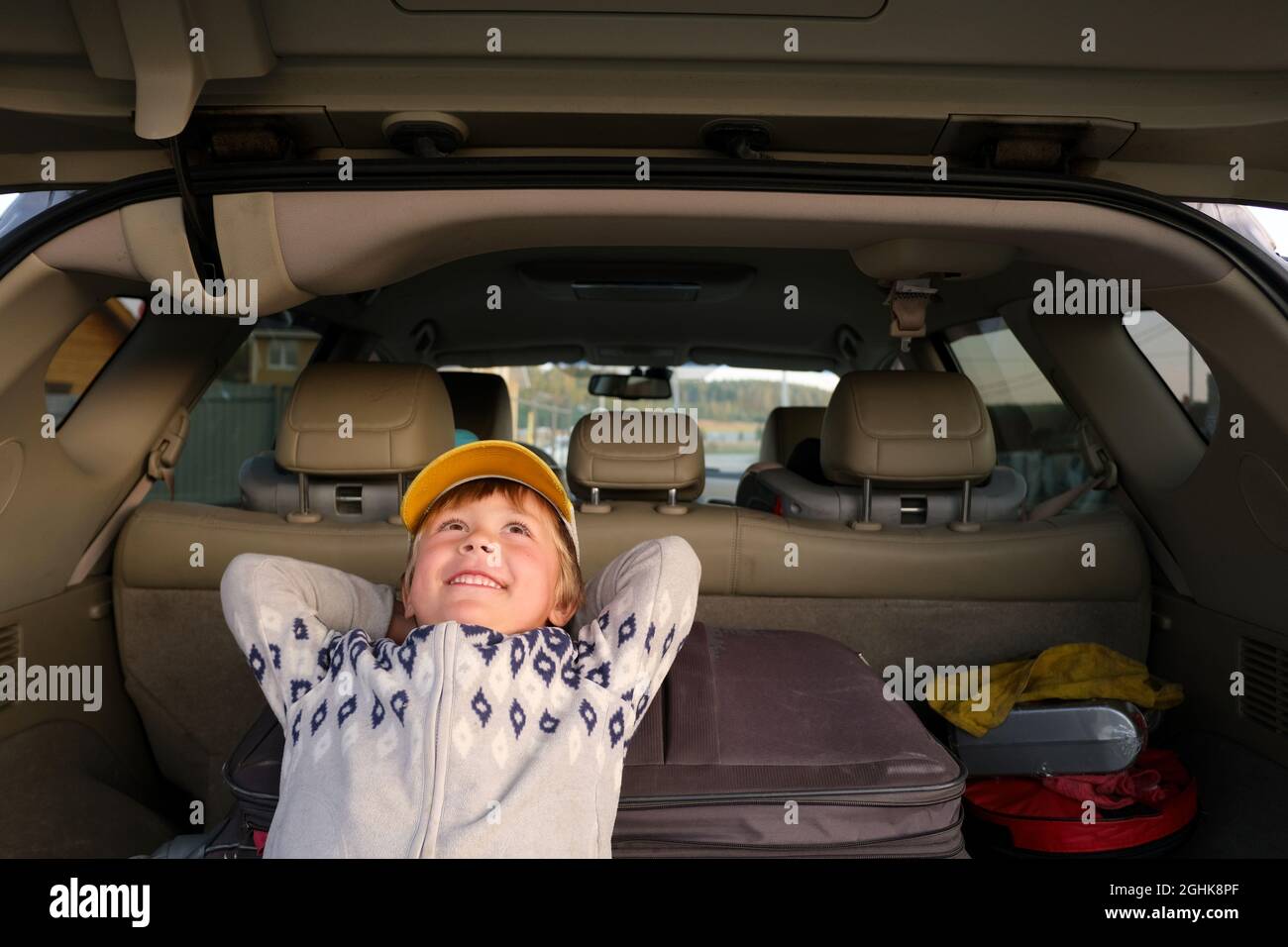 Verträumtes Kind in Mütze und Sweatshirt, das mit den Händen hinter dem Kopf im Kofferraum des Automobils liegt und während der Reise nach oben blickt Stockfoto Verträumtes Kind in Mütze und Sweatshirt, das mit den Händen hinter dem Kopf im Kofferraum des Automobils liegt und während der Reise nach oben blickt Stockfoto