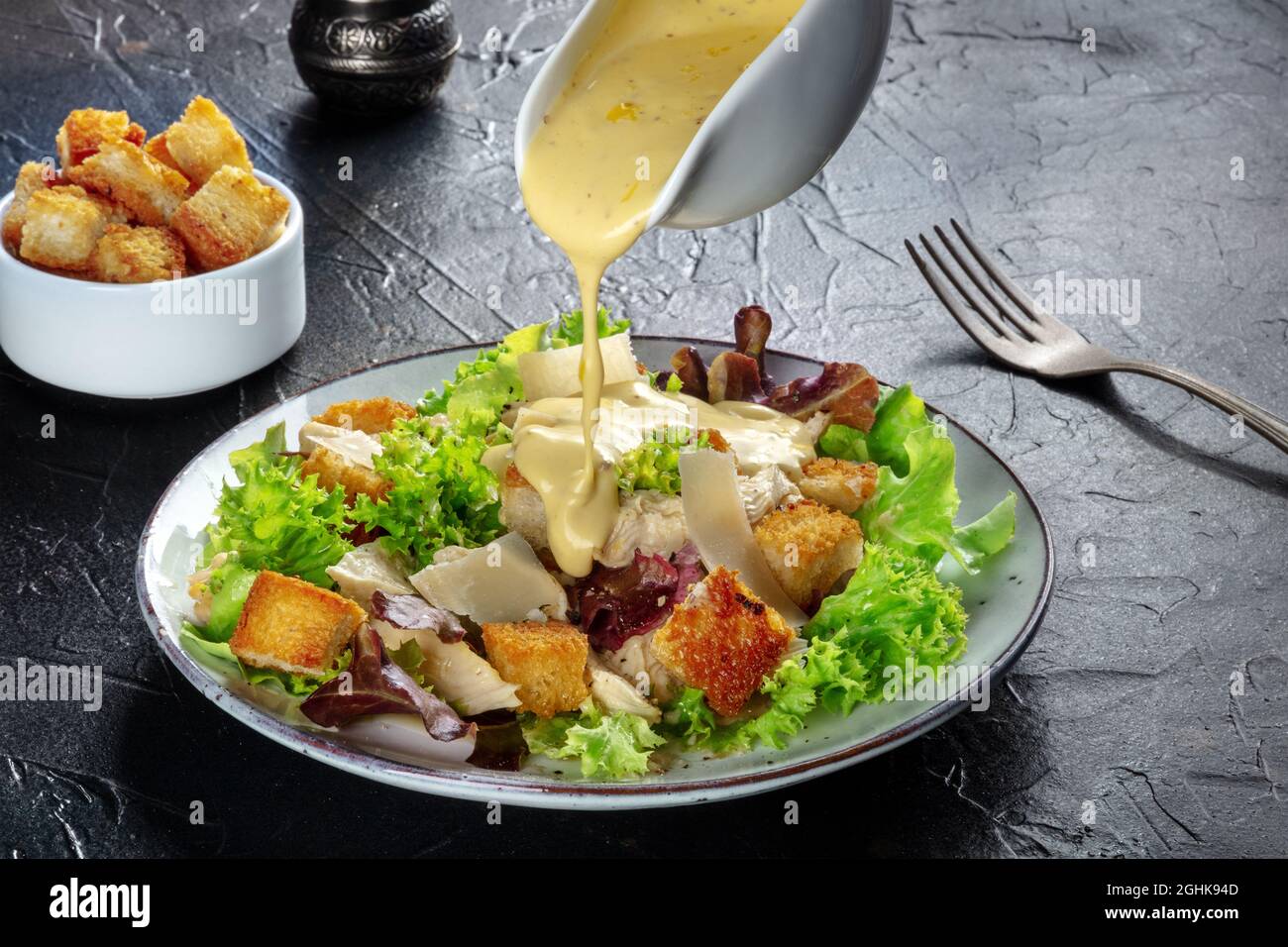 Chicken Caesar Salat mit dem klassischen Dressing, das gegossen wird, Croutons und Pfeffer auf schwarzem Hintergrund Stockfoto