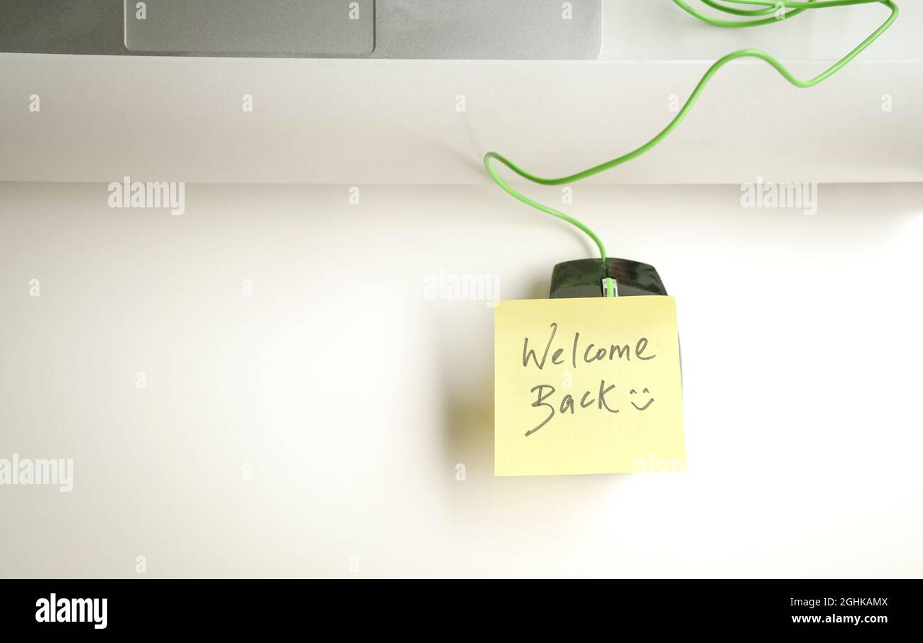 Willkommen zurück mit einem Smiley, der auf einem gelben Papierstock auf der Computermaus auf dem Bürotisch steht. Stockfoto