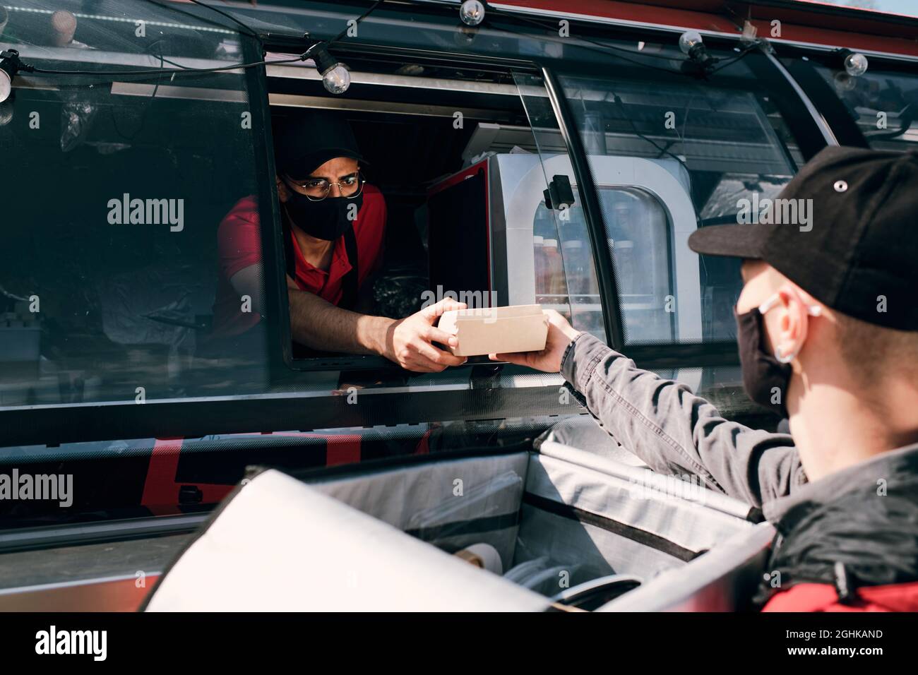 Junge Verkäuferin in Schutzmaske, die verpackte Schachtel mit Fast Food an das Personal des Lieferservice übergibt Stockfoto