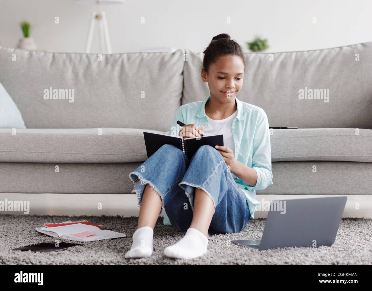 Lächelnde, afroamerikanische Schülerin studiert aus der Ferne, schreibt im Notebook, sitzt auf dem Boden und schaut auf den Laptop Stockfoto