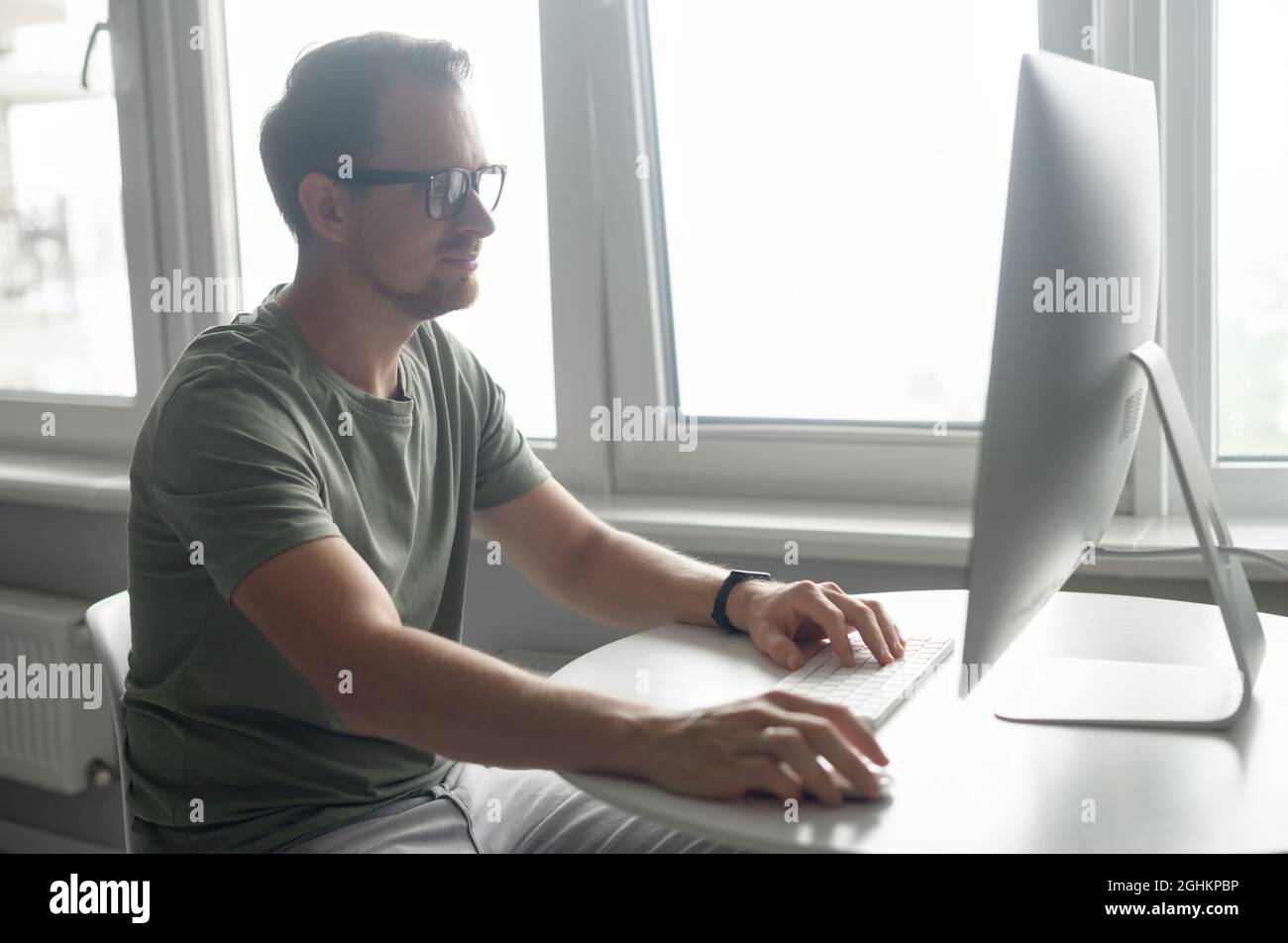 Schöner junger Hipster-Freiberufler mit Bart in Brille, der am Tisch sitzt und auf den PC-Monitor schaut, lächelnder Kerl, der gerne von zu Hause aus arbeitet, Nachrichten tippt und E-Mails beantwortet Stockfoto