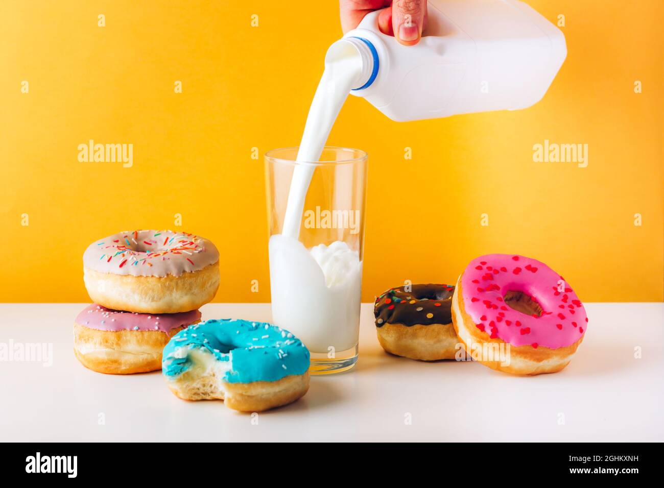 Bunte Donuts mit süßer Marmelade und Erdnussbutterfüllung auf grauem Tisch. Mann Hand gießt Pflanzenmilch auf das Glas. Vorderansicht gelb Stockfoto