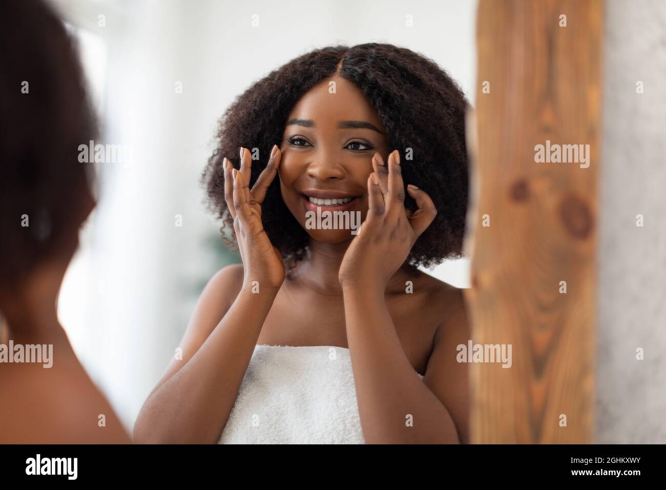 Charmante schwarze Dame lächelt sich im Spiegel an, überprüft die ersten Falten und bewundert die weiche Haut nach der Gesichtscreme zu Hause Stockfoto