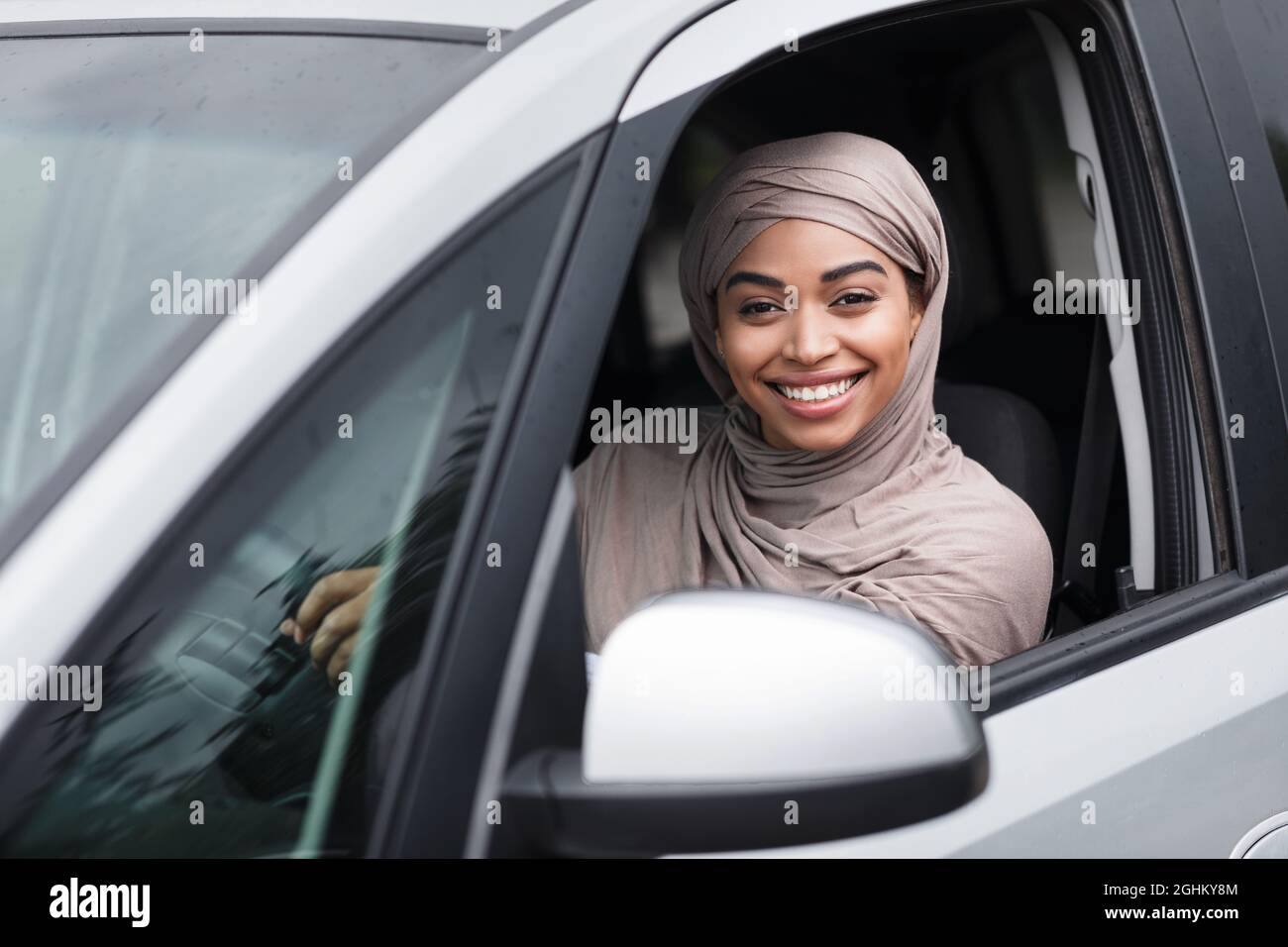 Fröhliche islamisch-afroamerikanische Fahrerin in Hijab sitzt im neuen Auto am Lenkrad und schaut auf die Kamera Stockfoto