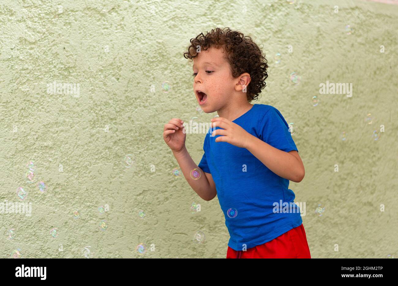 Lockiger Junge mit Sommersprossen, die im Hinterhof mit Seifenblasen spielen. Stockfoto