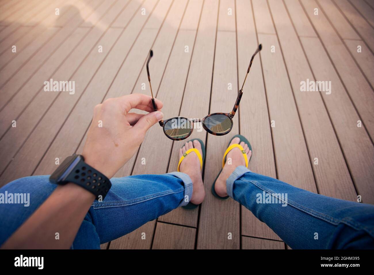 Selbstfoto der Hand Holding Brille mit Flips Flops Apple Watch auf dem Terrassenboden mit aufziehender Jeans Stockfoto