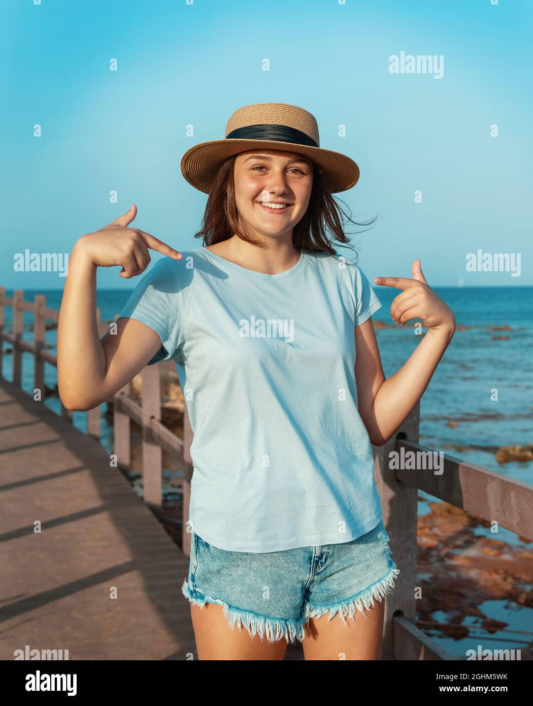 Lächelndes Teenager-Mädchen, das auf einem hölzernen Bürgersteig am Meer in Strohhut, hellblauem T-Shirt und mit dem Blick darauf bleibt. T-Shirt-Modell Stockfoto
