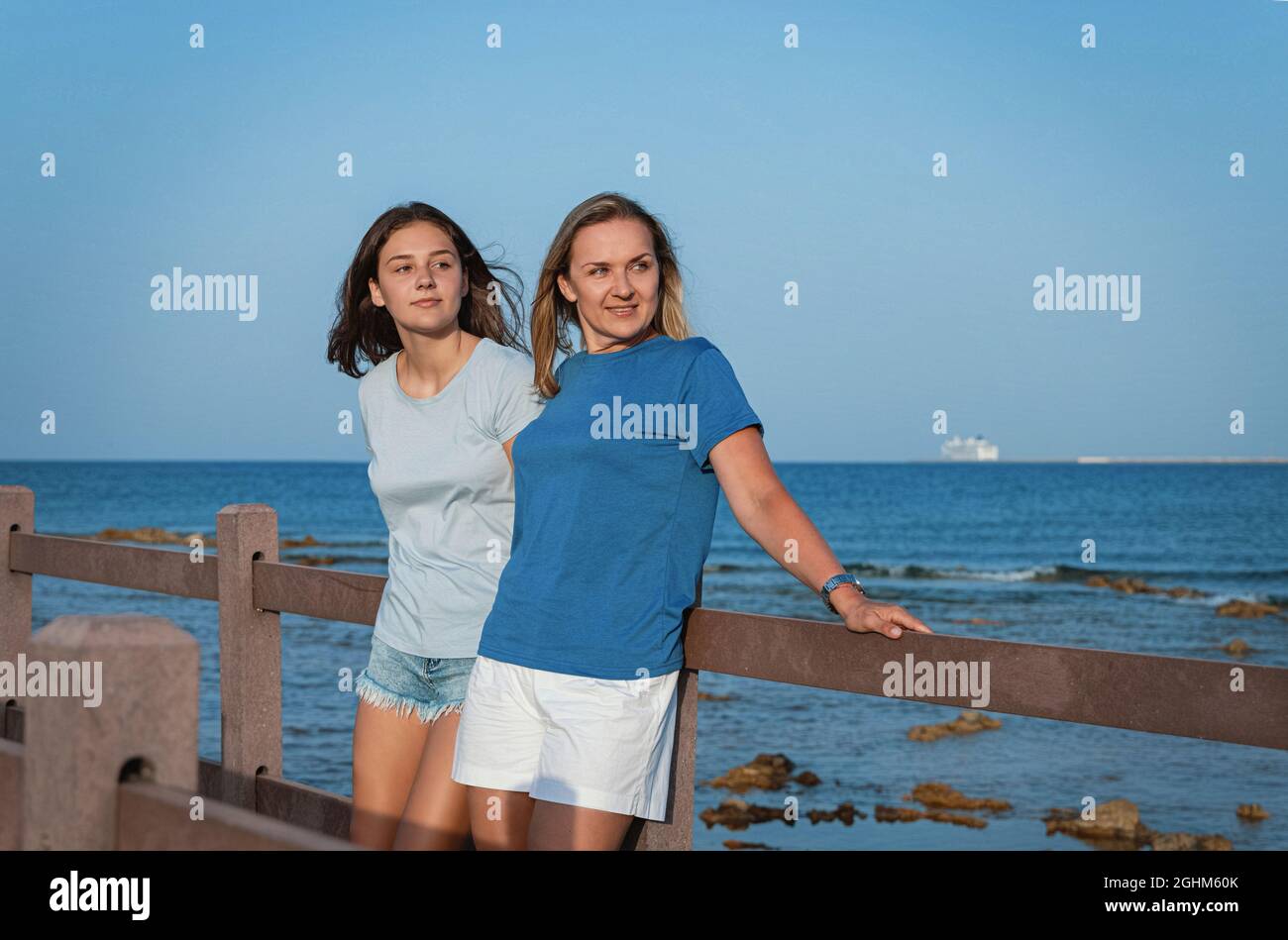 Mutter und Tochter stehen bei Sonnenuntergang auf einem hölzernen Bürgersteig am Meer. Eine Frau mittleren Alters und ein Mädchen im Teenageralter stehen nebeneinander und tragen ein blaues T-sh Stockfoto