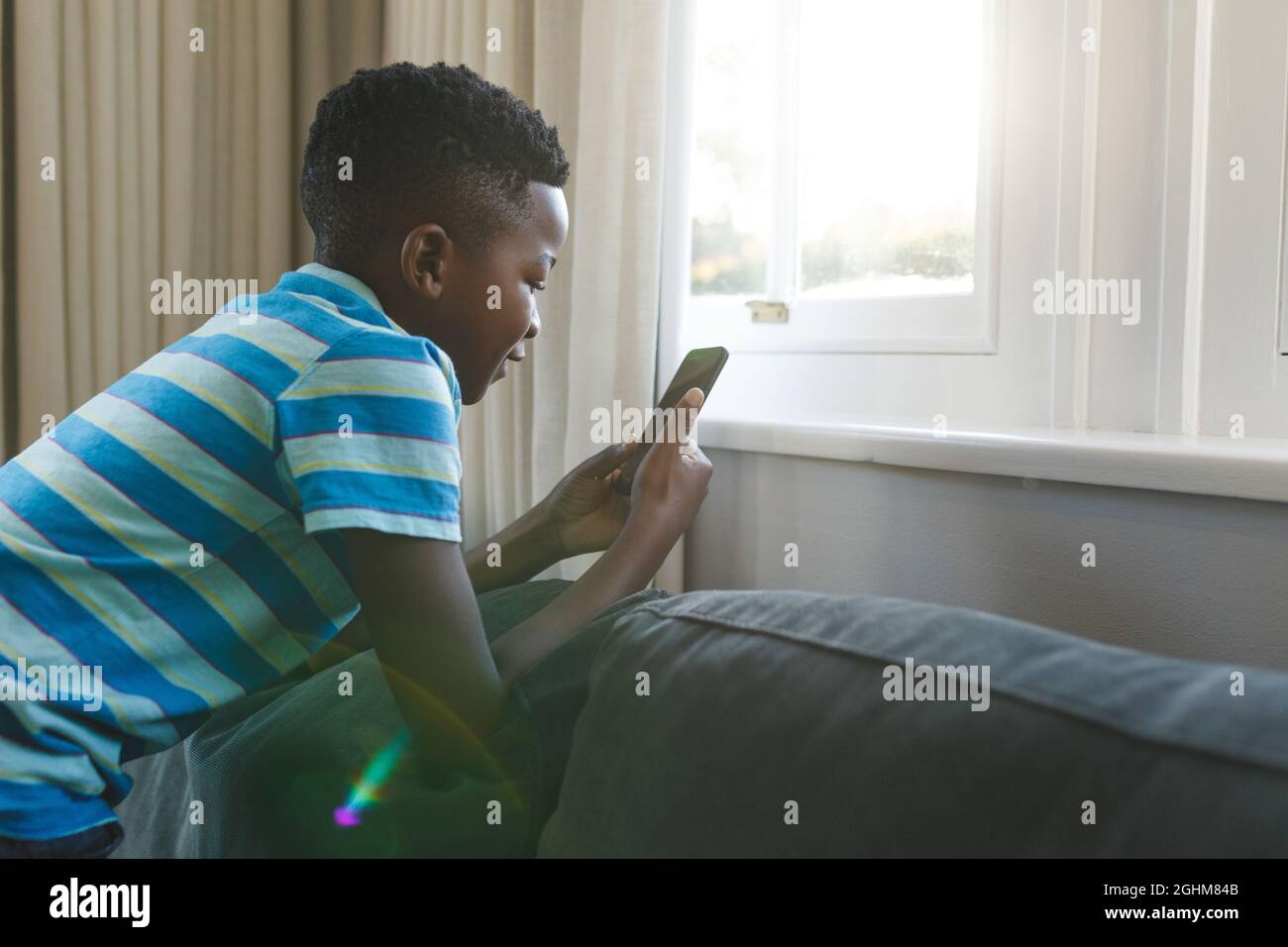 Glücklicher afroamerikanischer Junge, der mit dem Smartphone im Wohnzimmer auf der Couch kniet Stockfoto