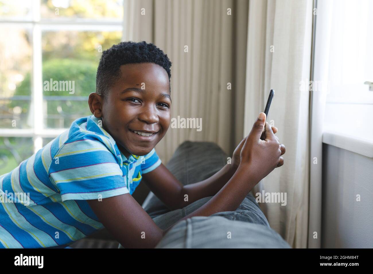 Porträt eines glücklichen afroamerikanischen Jungen, der mit dem Smartphone im Wohnzimmer auf der Couch kniet Stockfoto