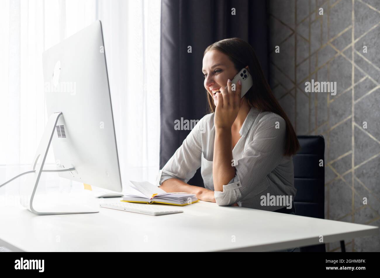 Eine charmante junge Frau telefoniert, eine Geschäftsfrau sitzt entspannt am Schreibtisch vor einem trendigen Computer, lacht und chattet auf dem Smartphone, während sie eine Pause vom Fernarbeiten macht Stockfoto