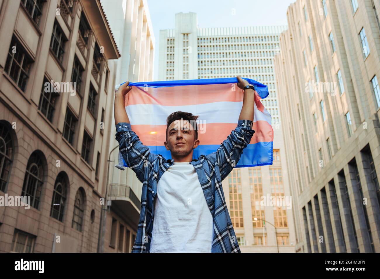 Transman, der die Transgender-Flagge im Freien aufhebt. Selbstbewusster junger Transgender-Mann, der den Stolz der Homosexuellen in der Stadt feiert. Junger Mann, der nicht dem Geschlecht entspricht, stan Stockfoto