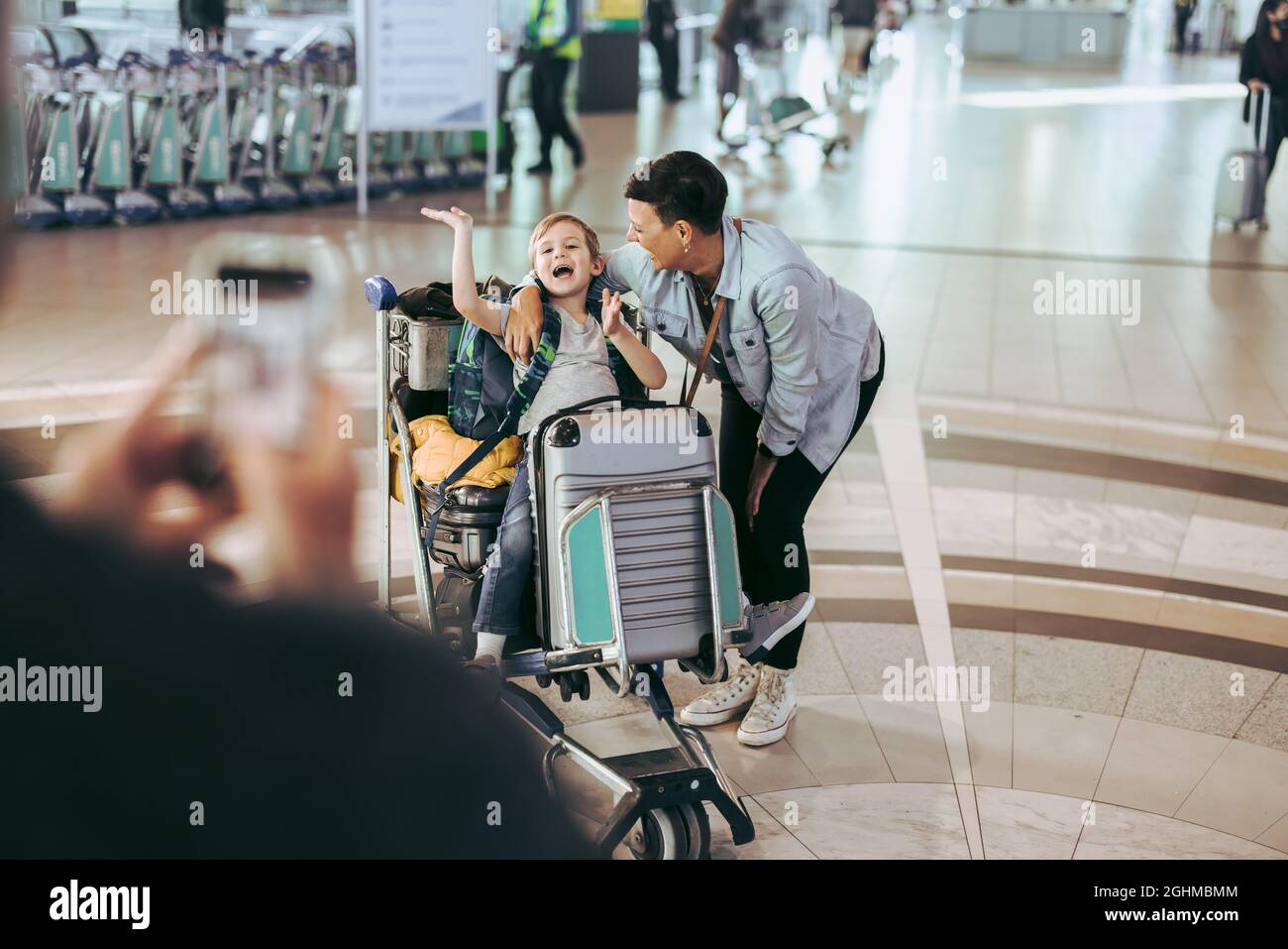 Mutter mit Sohn sitzt auf Gepäckwagen, während Vater auf Bilder klickt. Vater fotografiert Frau und Sohn am Flughafen. Stockfoto