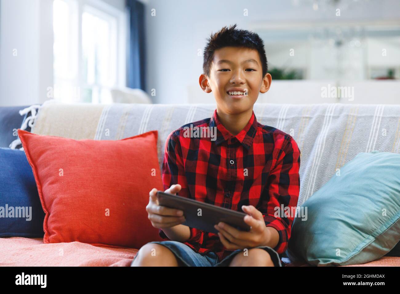 Porträt eines lächelnden asiatischen Jungen, der auf der Couch sitzt und ein Tablet benutzt Stockfoto