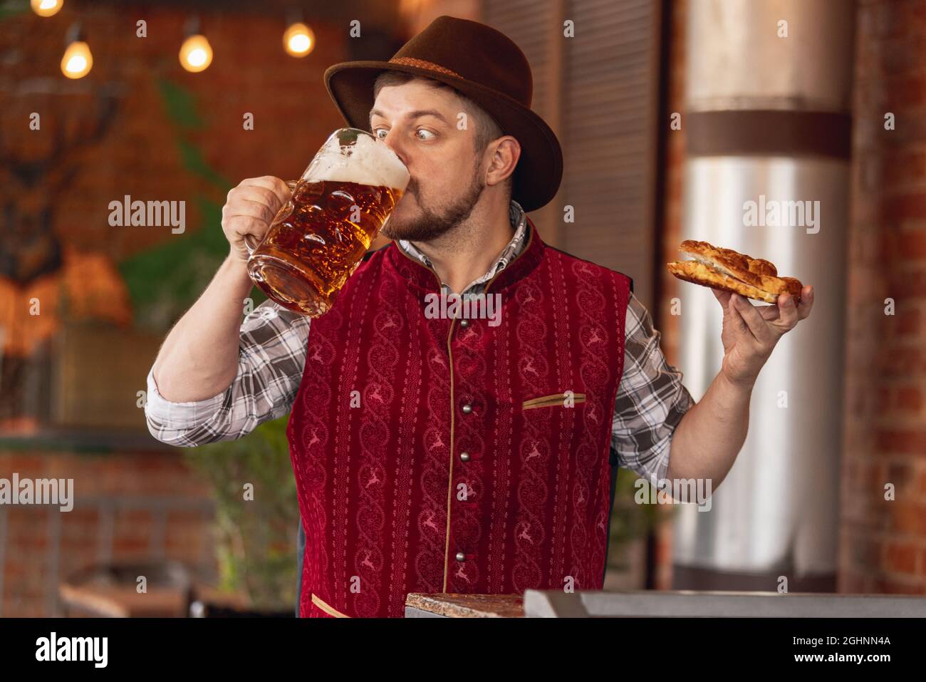 Porträt eines glücklichen bärtigen Mannes in traditioneller bayerischer Festtracht, Hut und roter Weste, der während des Bierfestes in Bar, Café, Pub Bier und Snack verkostet Stockfoto