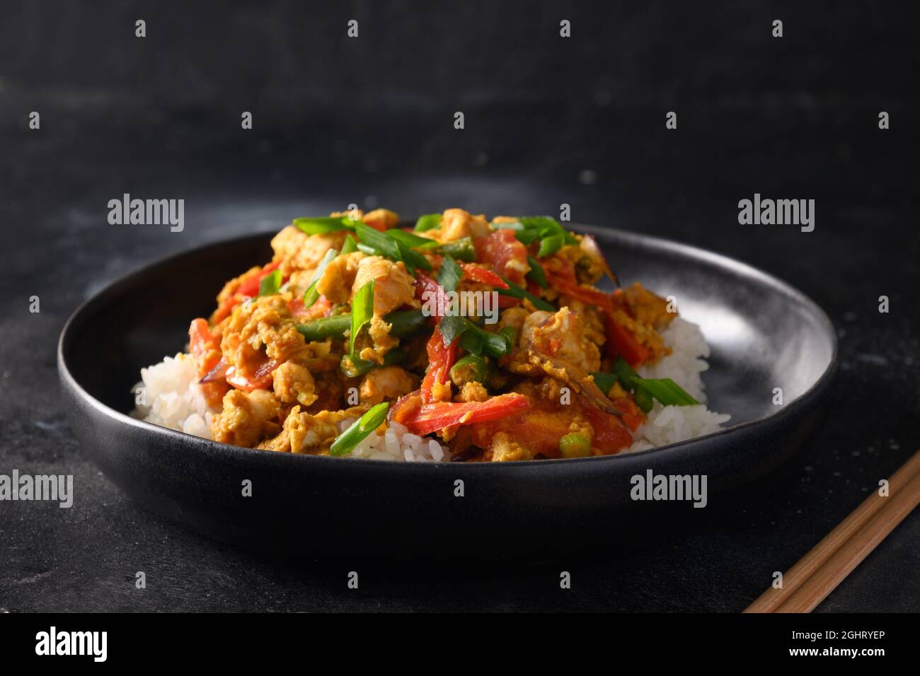 Thailändisches Gericht Gai Pad Pongali mit Huhn, Eiern, würziger, gelber thai-Paprikapaste, Tomaten, Reis in schwarzer Schüssel im Wok. Nahaufnahme. Traditionelle lokale Pause Stockfoto