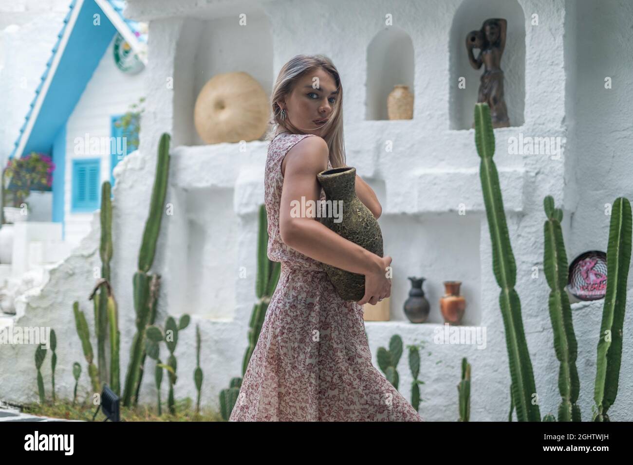 Porträt einer hübschen jungen Frau mit blonden langen Haaren in langen Kleid posiert mit alten Vase in der Hand. Weiße Designer-Wand mit Kakteen und Statuen Stockfoto