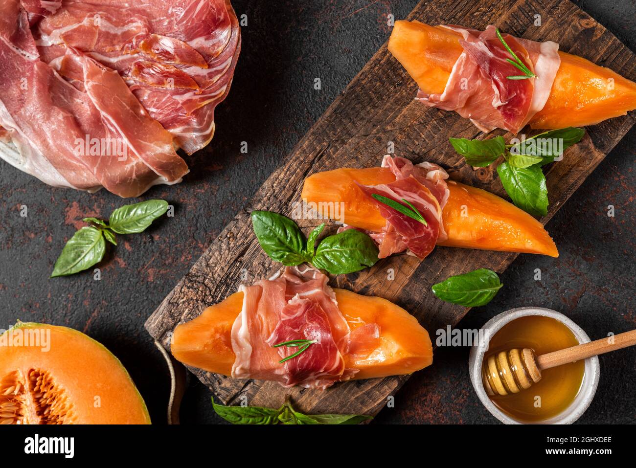 Prosciutto-Schinken mit Melone-Cantaloupe-Scheiben, Honig und Basilikum auf Schneidebrett auf dunklem Hintergrund. Italienische Vorspeise. Draufsicht. Gesunde Ernährung Stockfoto