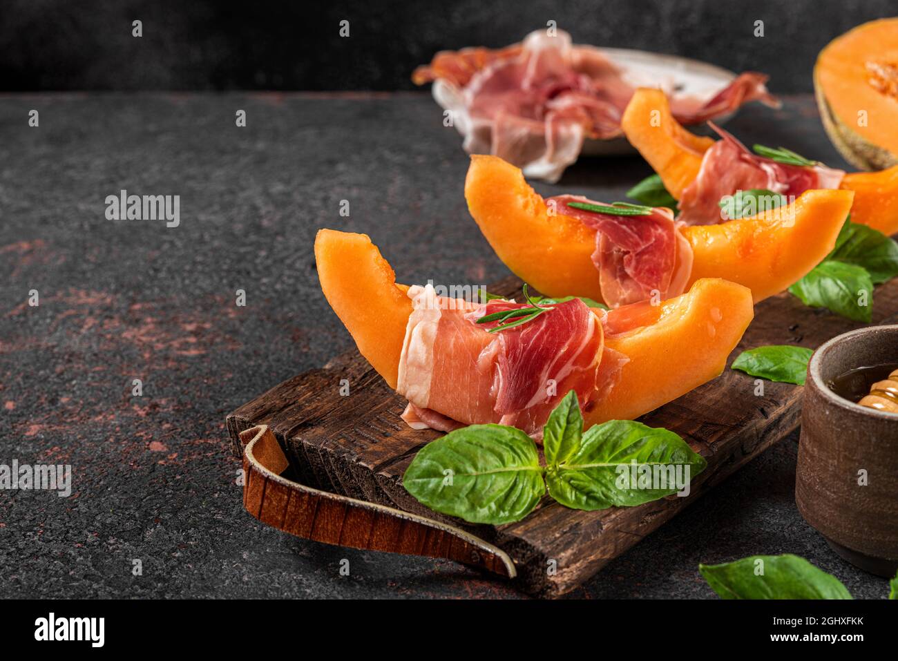 Prosciutto-Schinken mit Melone-Cantaloupe-Scheiben, Honig und Basilikum auf Schneidebrett auf dunklem Hintergrund. Italienische Vorspeise. Gesunde Ernährung Stockfoto