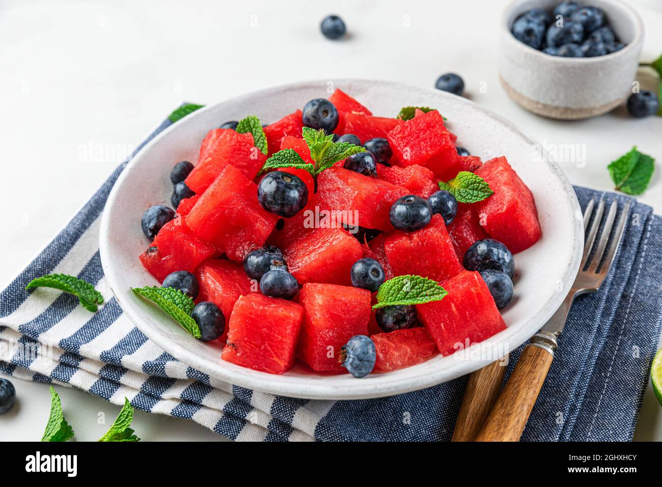 Wassermelonen- und Heidelbeer-Obstsalat mit Minze und Limettensaft auf einem Teller über Serviette auf weißem Hintergrund. Nahaufnahme. Erfrischendes Sommeressen Stockfoto