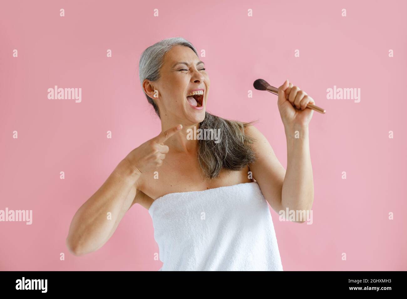 Aufgeregt mittleren Alters asiatische Modell mit Frottee Handtuch gewickelt singt auf rosa Hintergrund Stockfoto