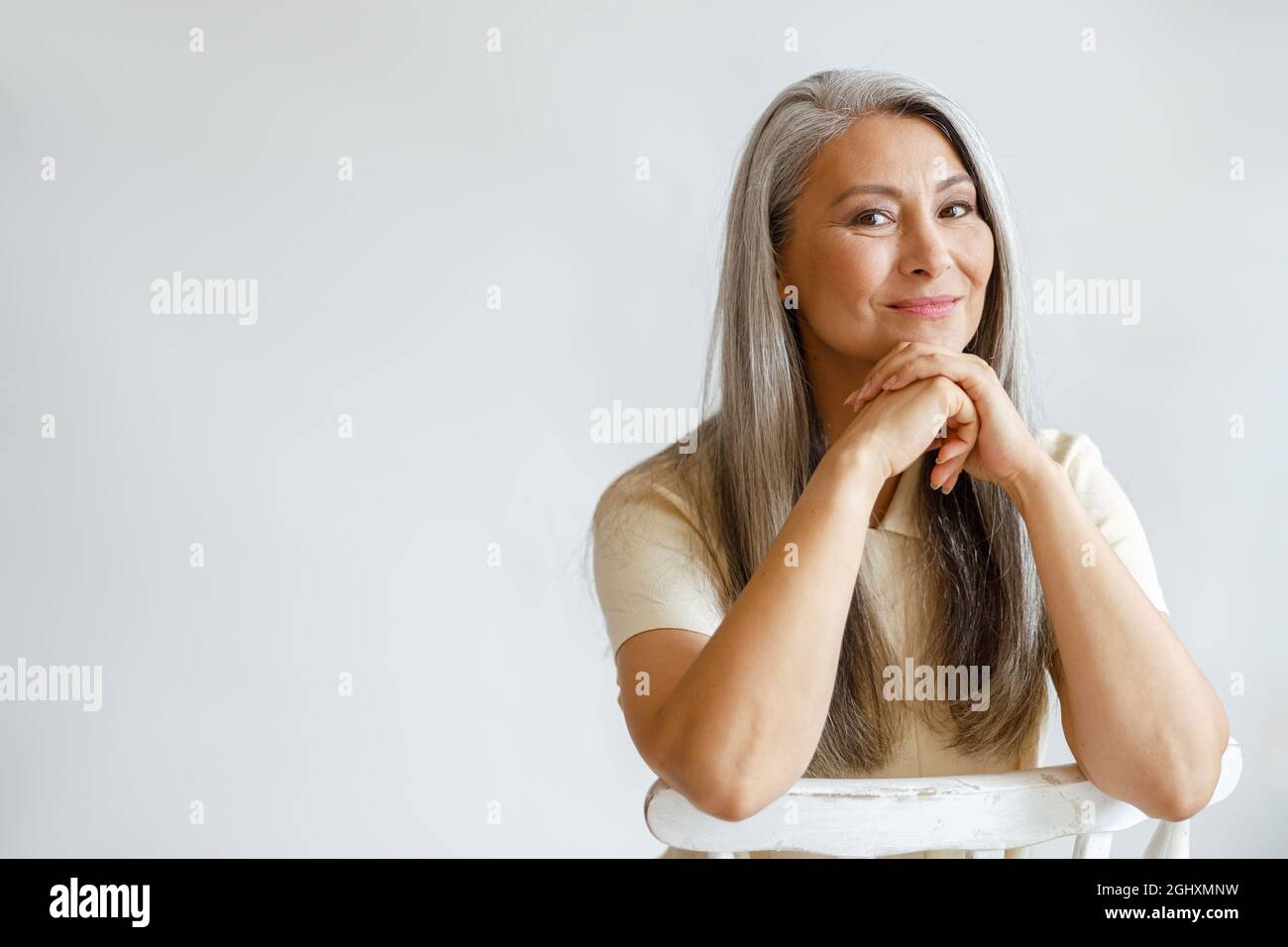 Glückliche Frau mittleren Alters mit silbernen Haaren sitzt auf den Händen auf hellem Hintergrund gelehnt Stockfoto
