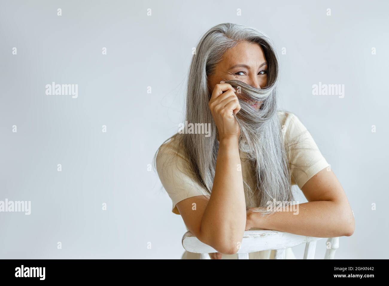 Positive asiatische Dame mittleren Alters verwendet heidselige Haare als Niqab, der im Studio auf einem Stuhl sitzt Stockfoto