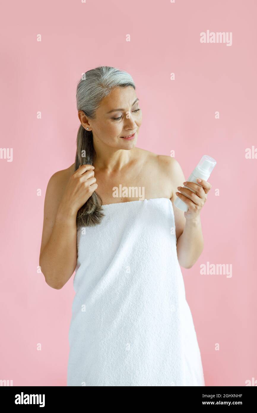 Silberhaarige asiatische Frau mittleren Alters sieht Körperpflegeprodukt auf rosa Hintergrund Stockfoto
