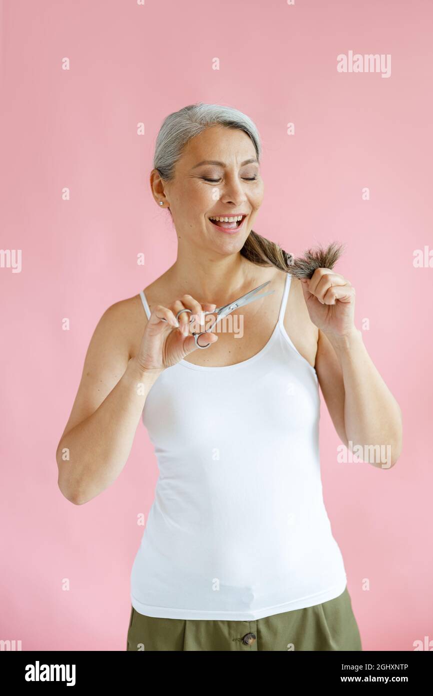 Positive asiatische Frau mittleren Alters schneidet gespaltene Enden von heidschem Haar mit einer Schere im Studio Stockfoto