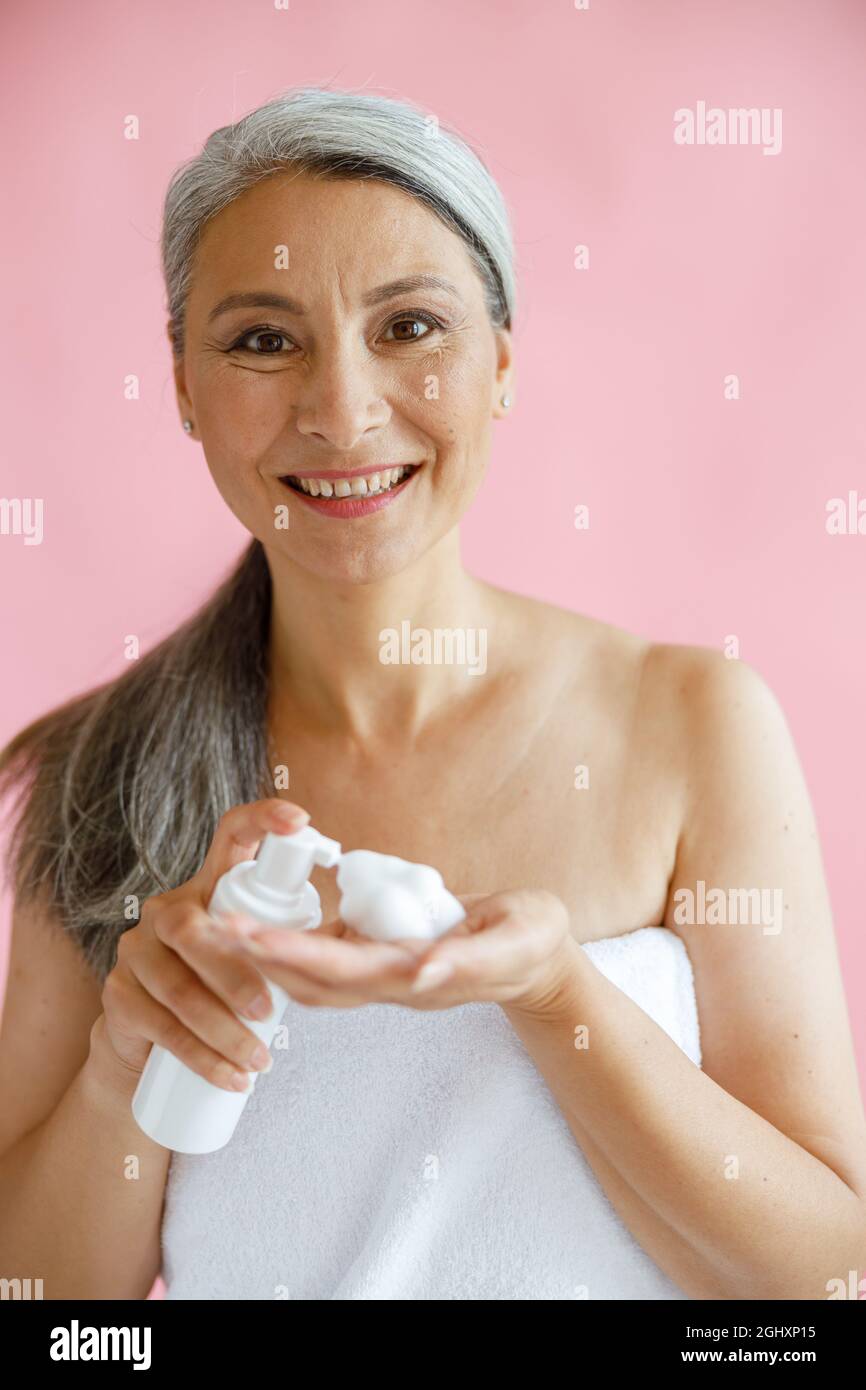 Positive asiatische Dame mittleren Alters, eingewickelt mit einem weichen Handtuch, hält Reinigungsschaum auf rosa Hintergrund Stockfoto