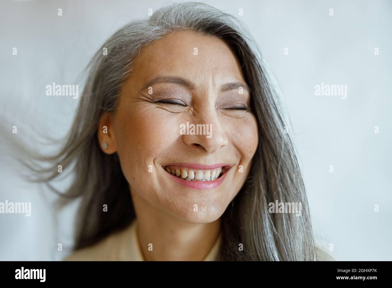 Fröhliches asiatisches Frauenmodell mittleren Alters mit fliegenden silbernen Haaren auf hellgrauem Hintergrund Stockfoto
