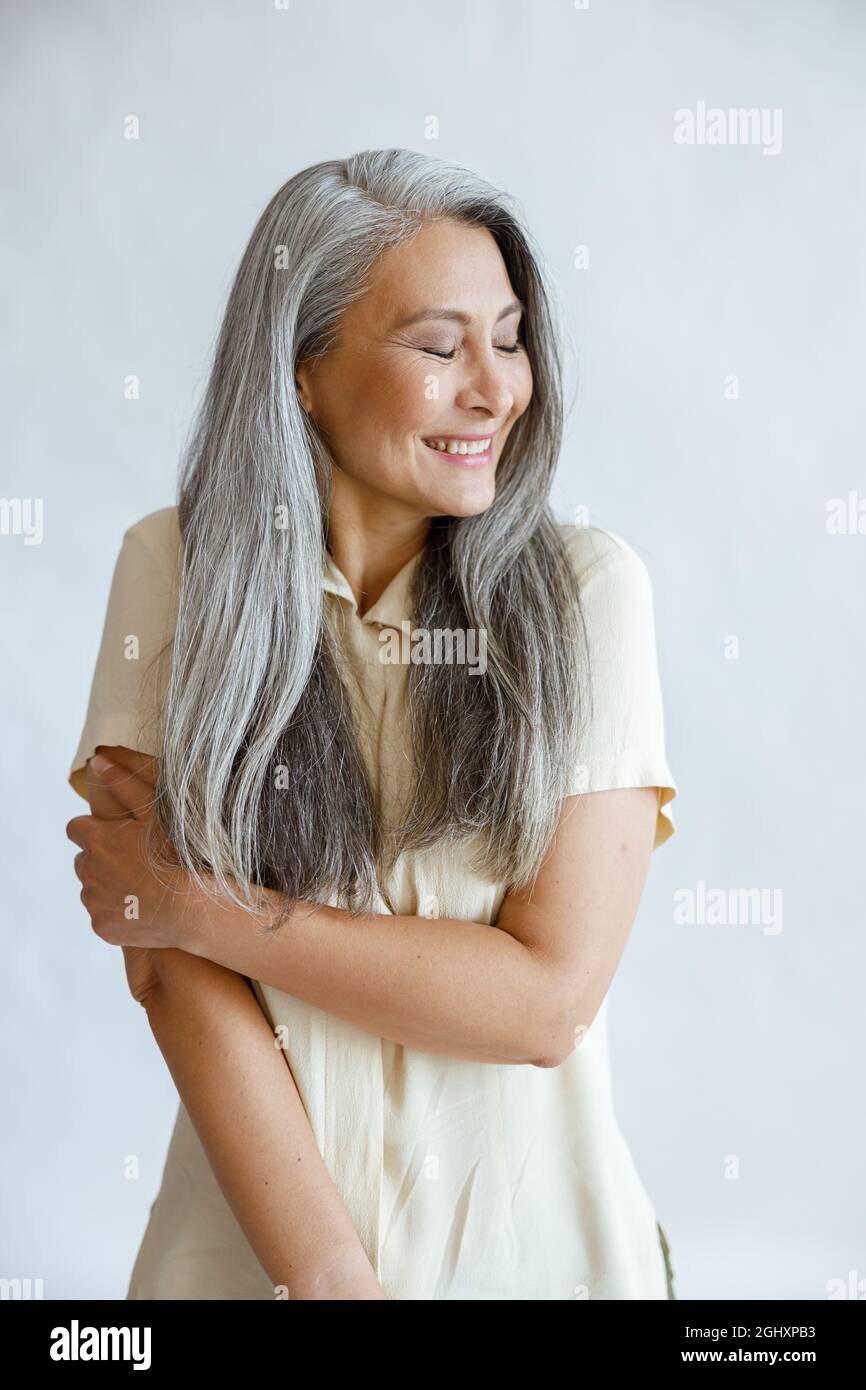 Scheue asiatische Dame mittleren Alters mit langen silbernen Haaren Posen auf hellgrauem Hintergrund Stockfoto