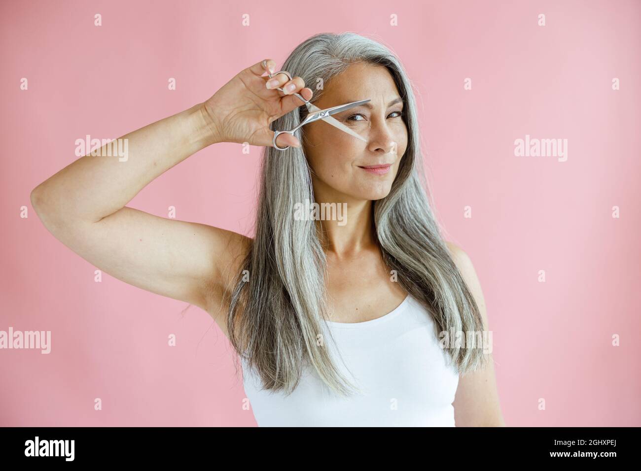 Schöne asiatische Dame mit grauen Haaren hält offene Schere in der Nähe des Gesichts auf rosa Hintergrund Stockfoto