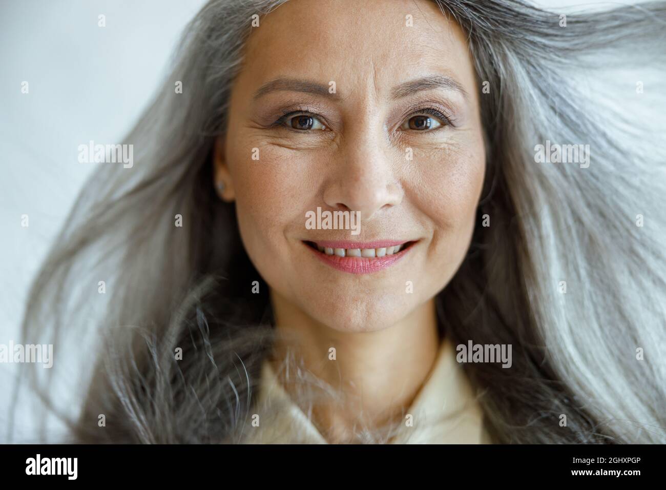 Eine Asiatin mittleren Alters mit fliegendem, grauem Haar blickt auf hellgrauen Hintergrund in die Kamera Stockfoto